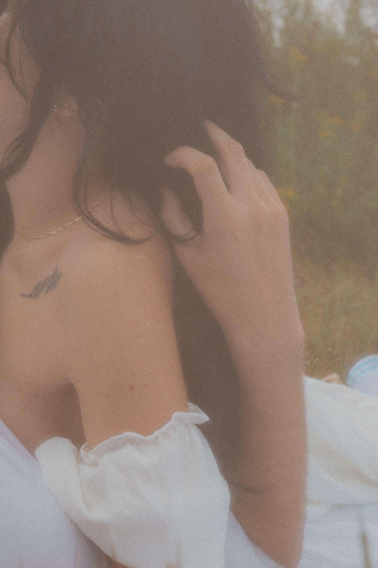 Close-up of woman's shoulder with delicate wing tattoo, wearing white off-shoulder dress in nature.