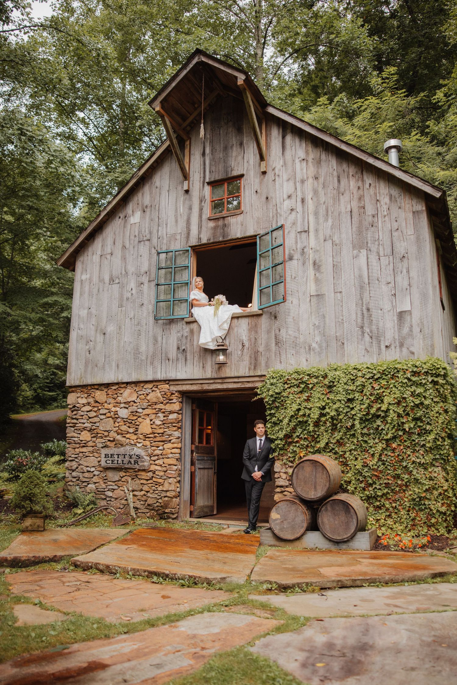 Bride sitting in barn window while groom stands below near wine barrels at rustic wedding venue.