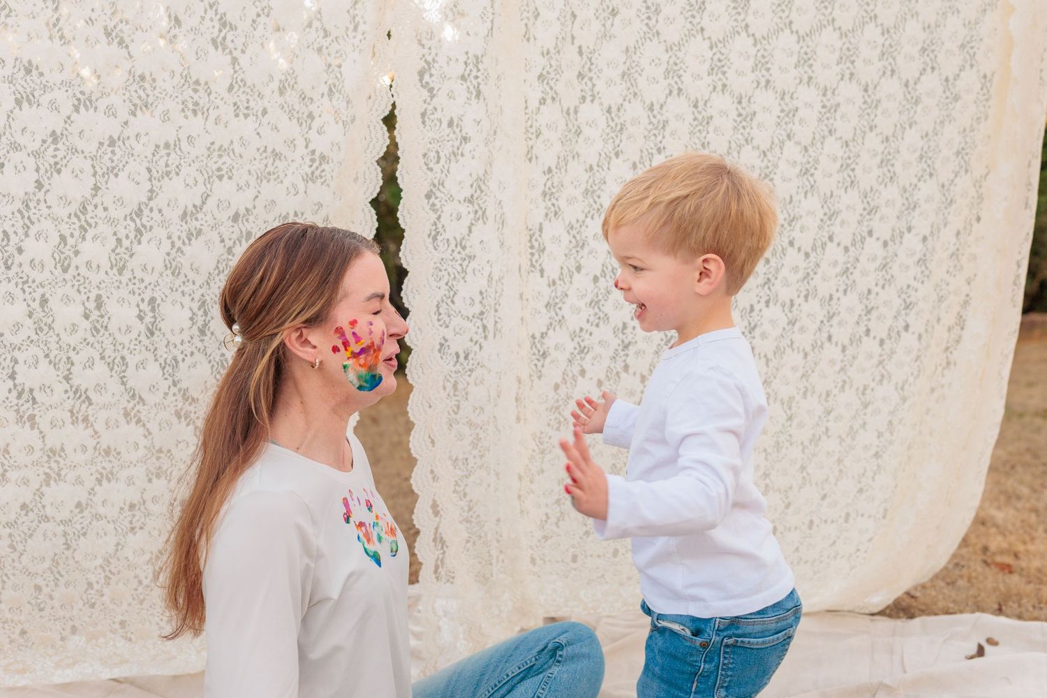 Woman wearing colorful face mask kneels beside laughing toddler boy in front of white lace backdrop outdoors.