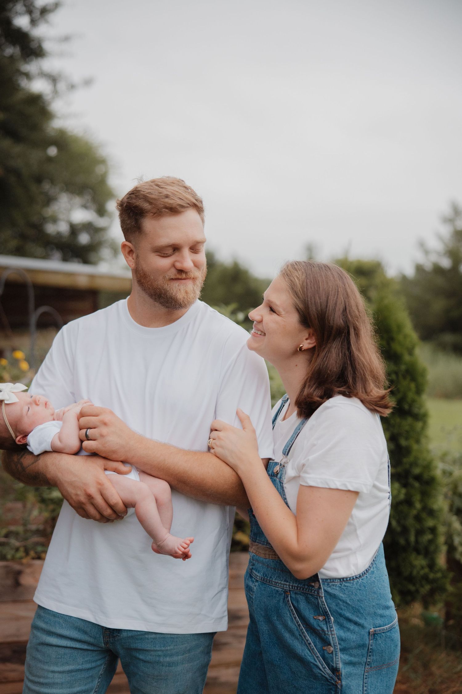 A young couple in white shirts smiles at each other while the man gently holds their newborn baby outdoors.