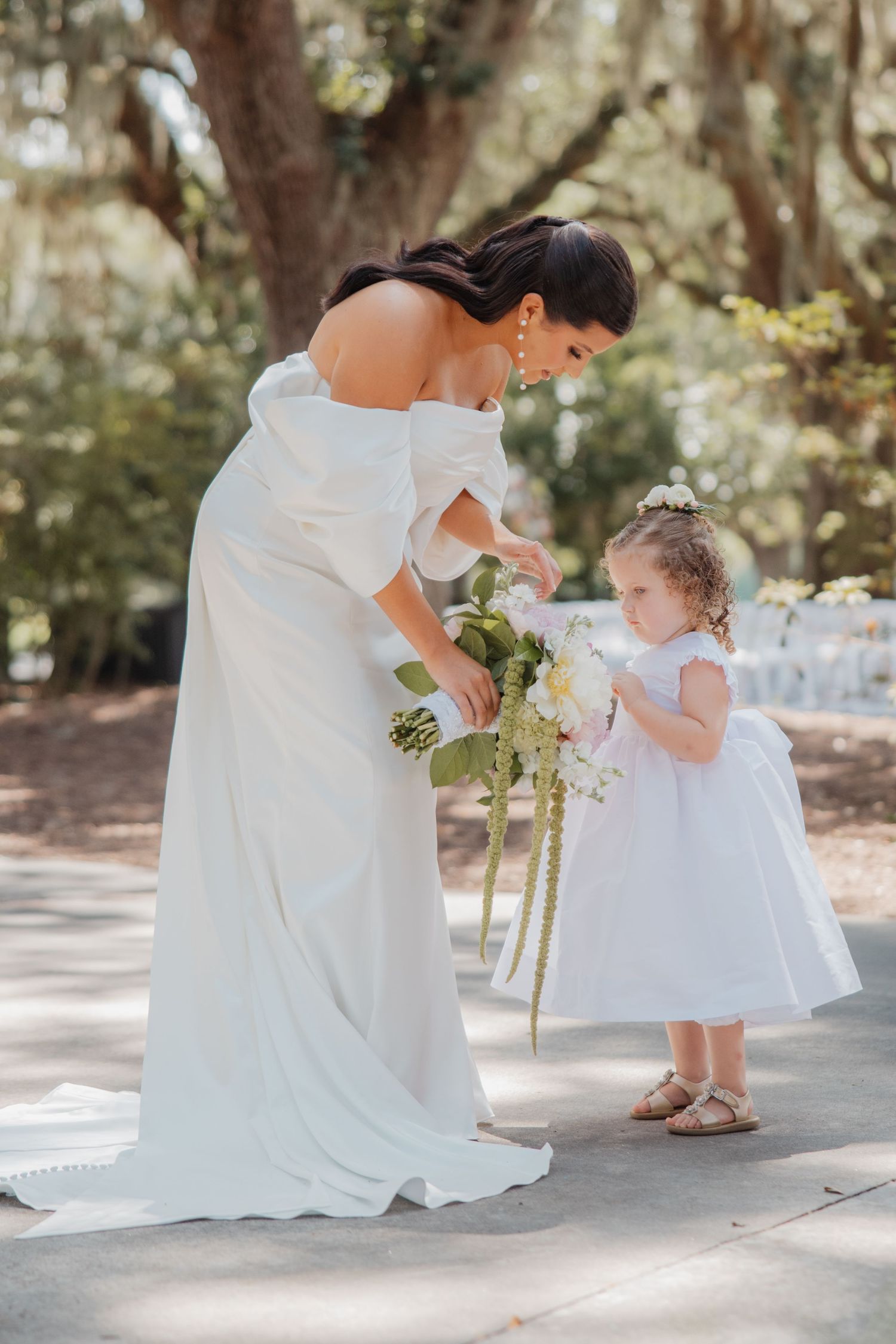 Bride in off-shoulder white gown bending down to share bouquet with flower girl in white dress outdoors.