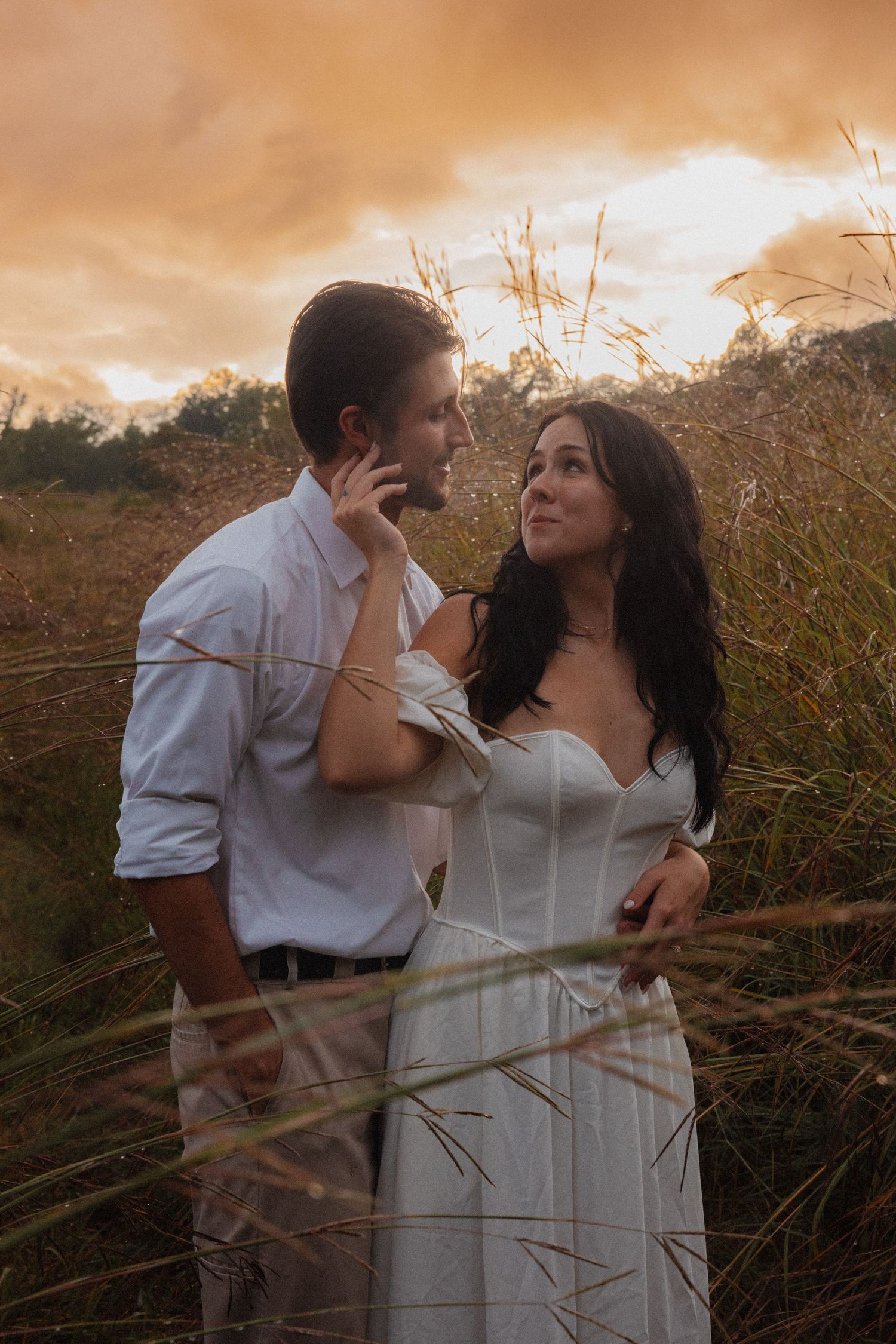 Couple in white embracing in tall golden grass at sunset with dramatic orange sky.