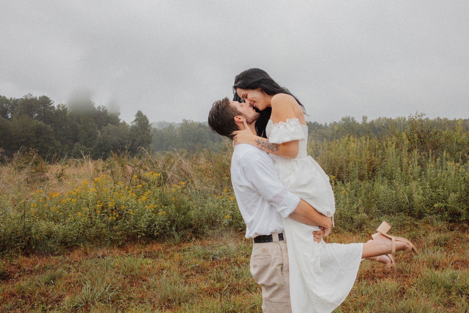 Couple sharing a romantic kiss in a misty field, man lifting woman in white off-shoulder dress outdoors.