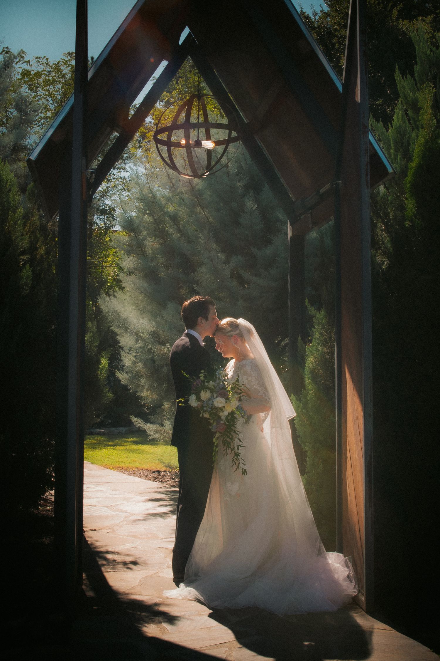 Bride and groom share a romantic moment under a wooden arch chapel entrance, surrounded by lush greenery in soft sunlight.