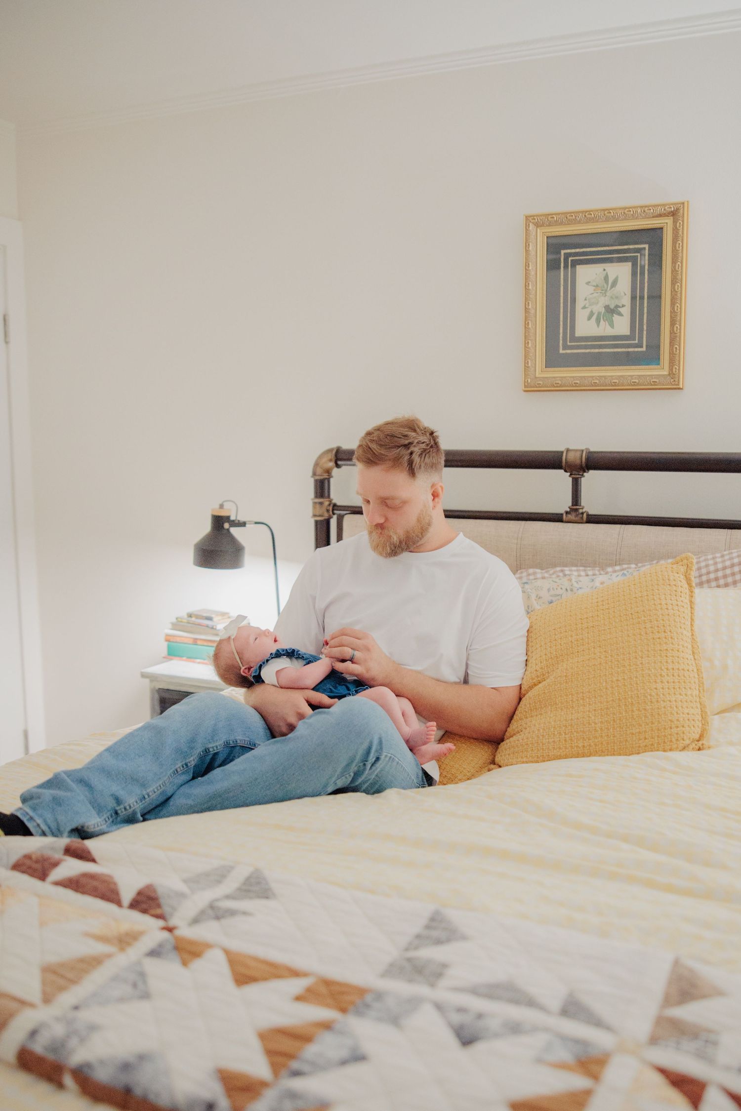 A father sits on a bed holding his newborn baby, surrounded by a colorful quilt and yellow pillows in a cozy bedroom.