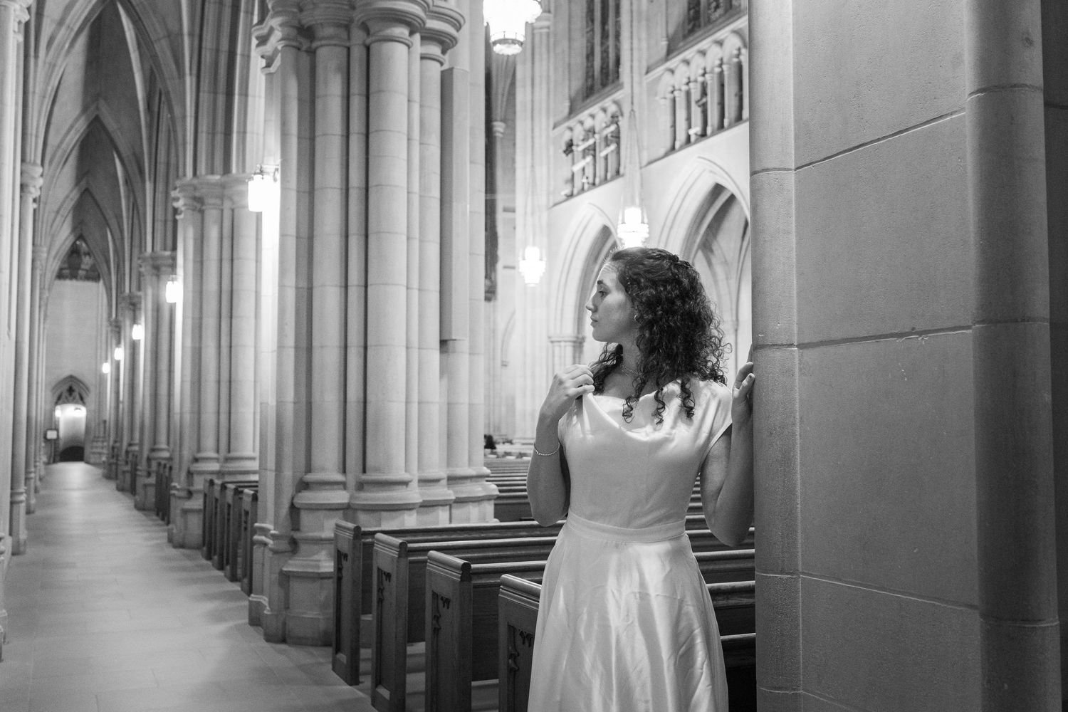 Black and white photo of a woman in white dress leaning against column in grand cathedral.