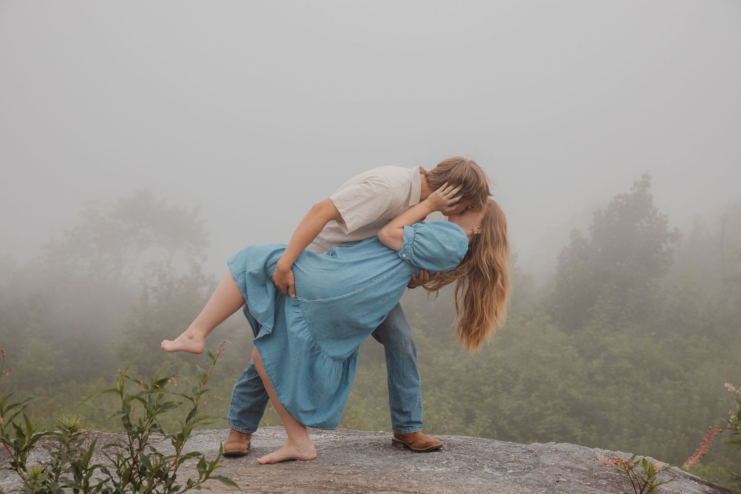 Couple sharing a romantic dip kiss on a misty mountain rock surrounded by foggy green trees.