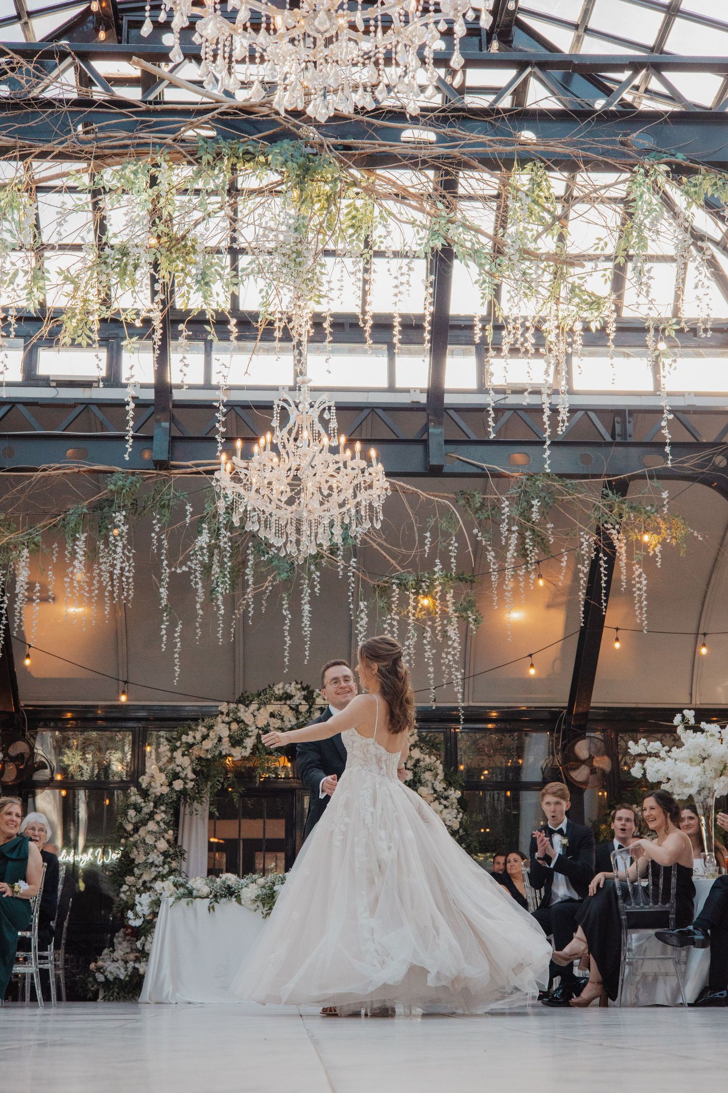Bride and groom sharing first dance under crystal chandeliers and hanging greenery in elegant venue.