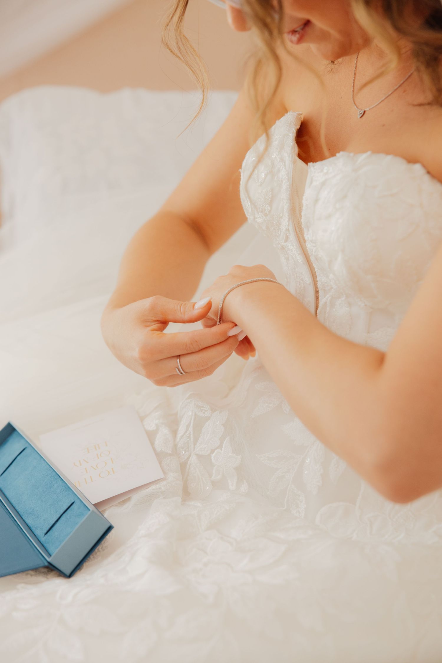 Bride in lace gown putting on crystal earrings beside a blue jewelry box on wedding day.