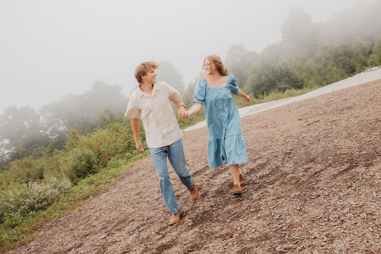 Young couple holding hands and laughing while walking on a foggy mountain trail with greenery.
