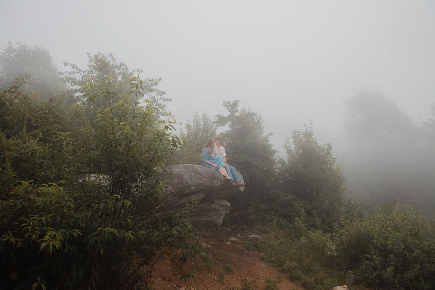 Woman in a blue dress sitting alone on a rock surrounded by misty green trees and fog.
