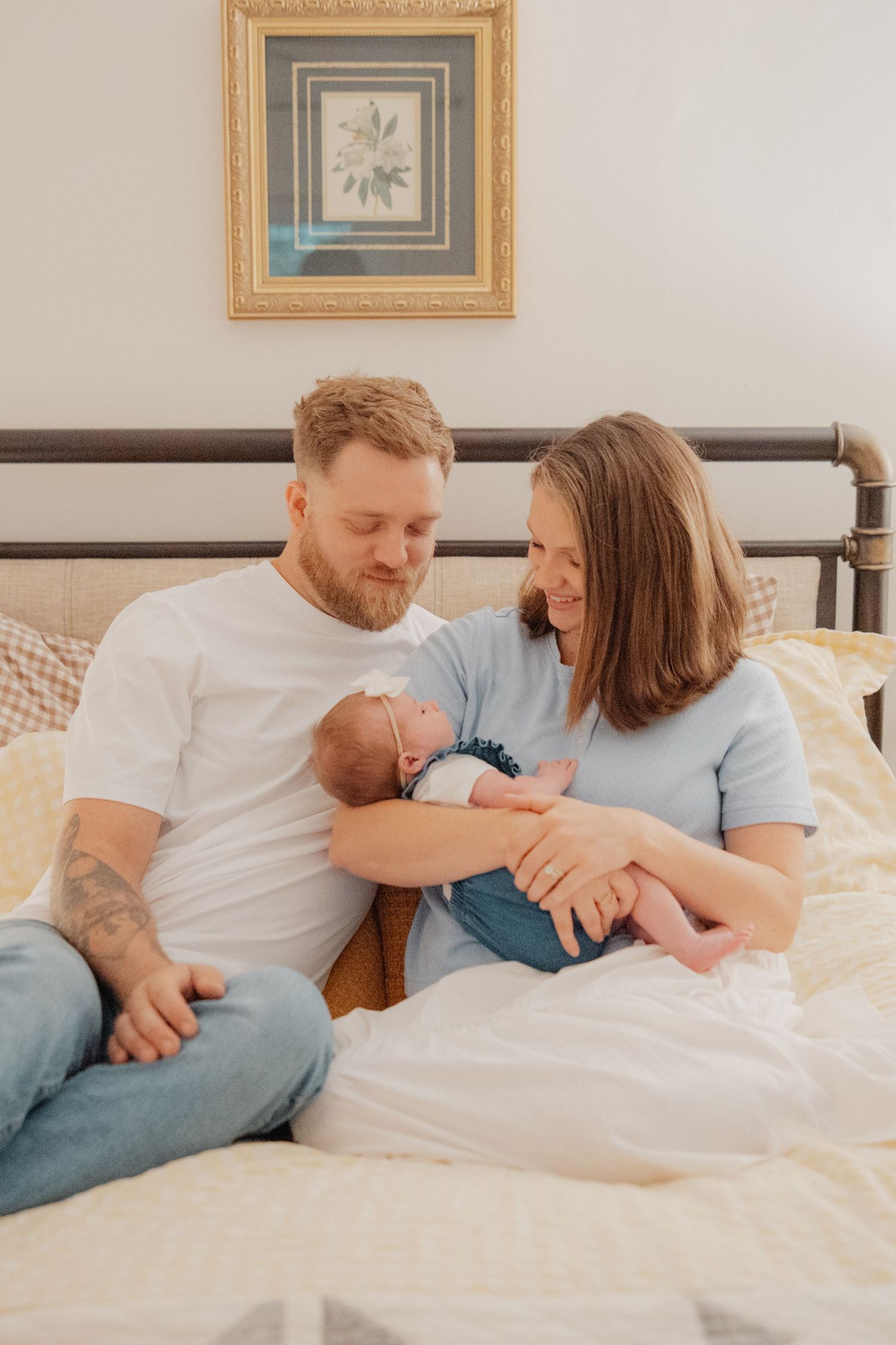 A couple sits on a bed smiling at their newborn baby, with a framed botanical print on the wall behind them.