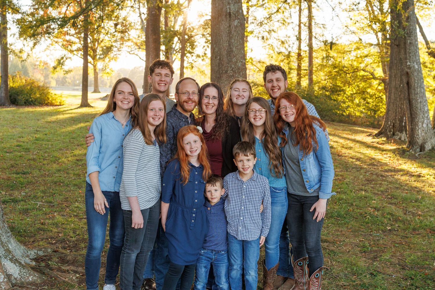 Large family group smiling outdoors in a sunlit park with tall trees and green grass in the background.