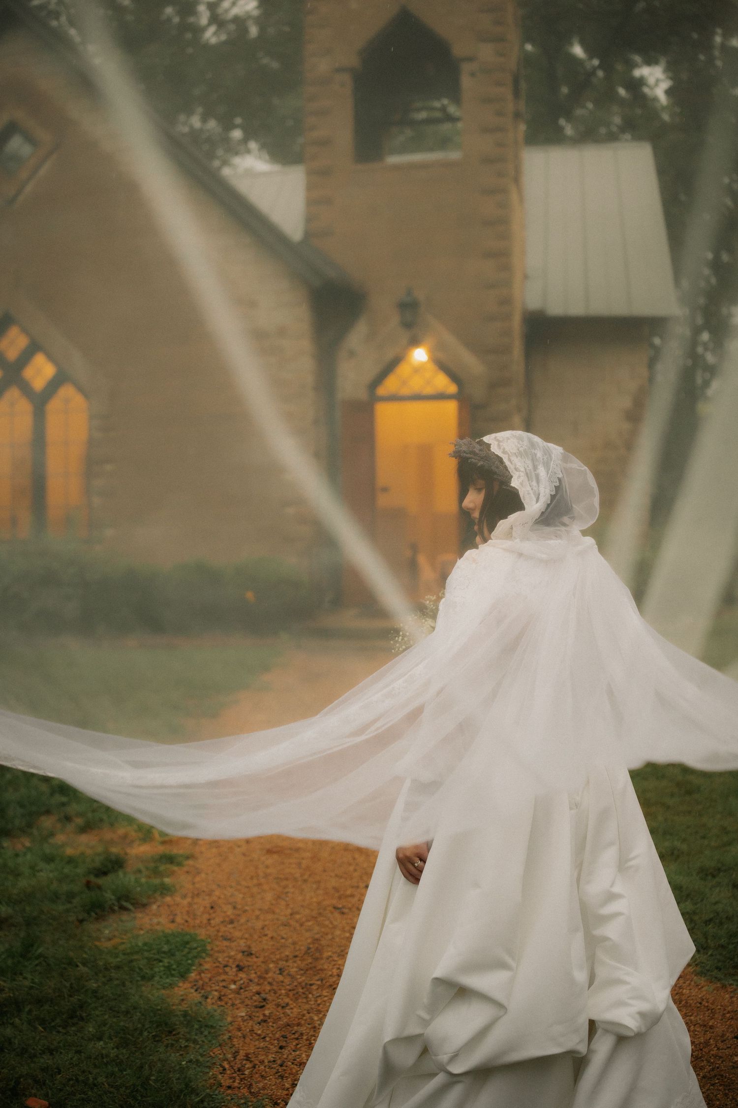 Bride in flowing white gown and veil walks toward a glowing stone chapel on a misty, atmospheric wedding day.