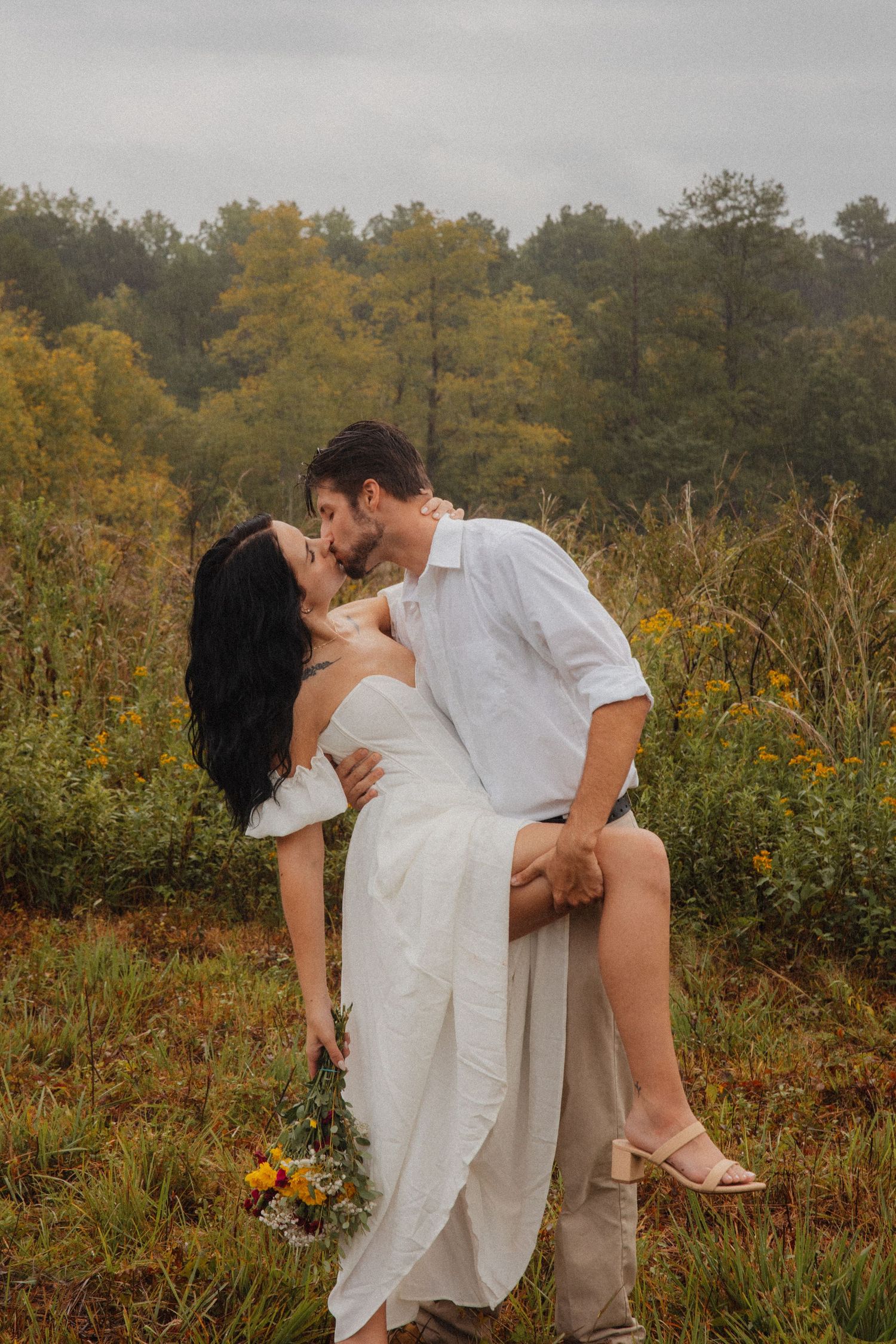 Couple sharing a romantic kiss outdoors, bride in white dress holding wildflower bouquet, groom in white shirt, autumn foliage background.