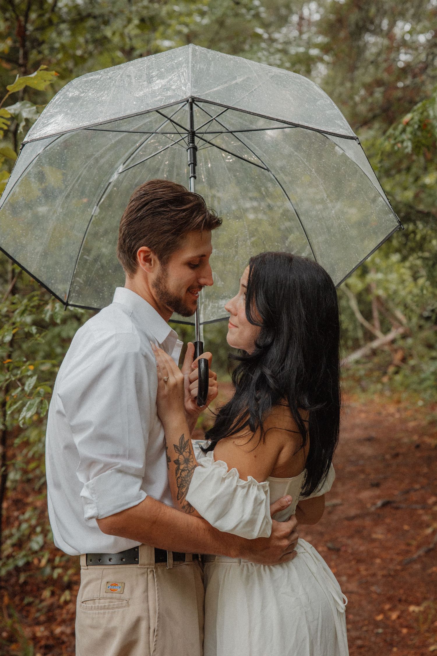 Couple embracing and smiling under a clear umbrella in a rainy green forest setting.
