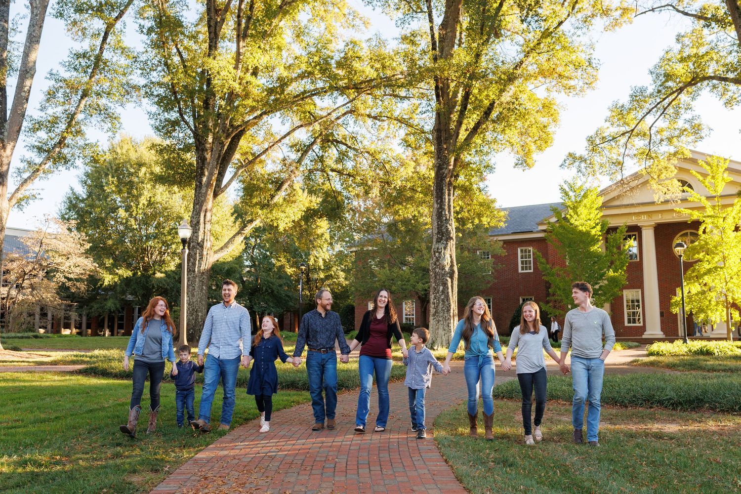 Large family holding hands walking on brick path surrounded by tall trees and red brick building in autumn sunlight.
