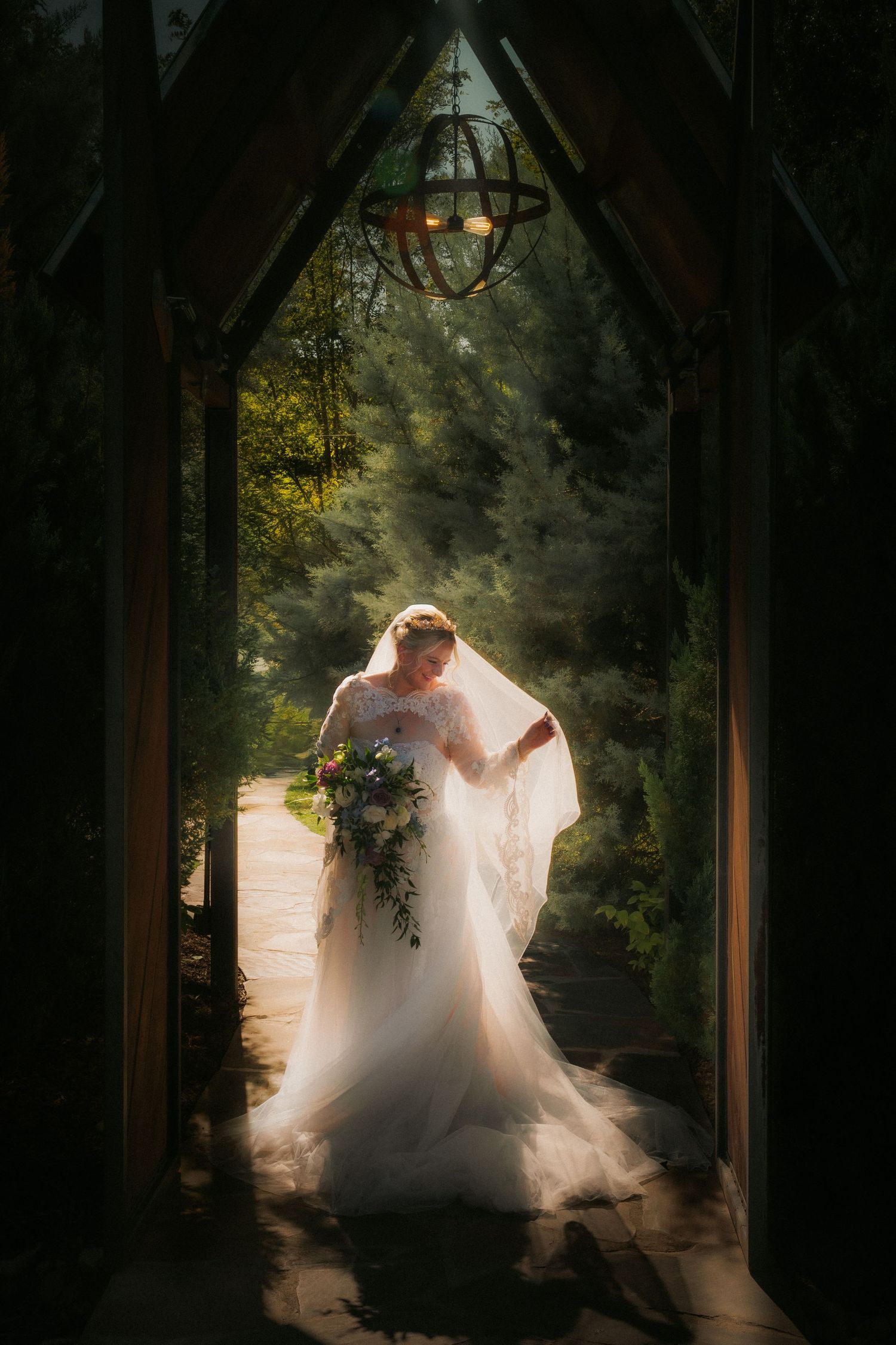 Bride in white gown and veil holding bouquet stands in sunlit garden archway with chandelier overhead.