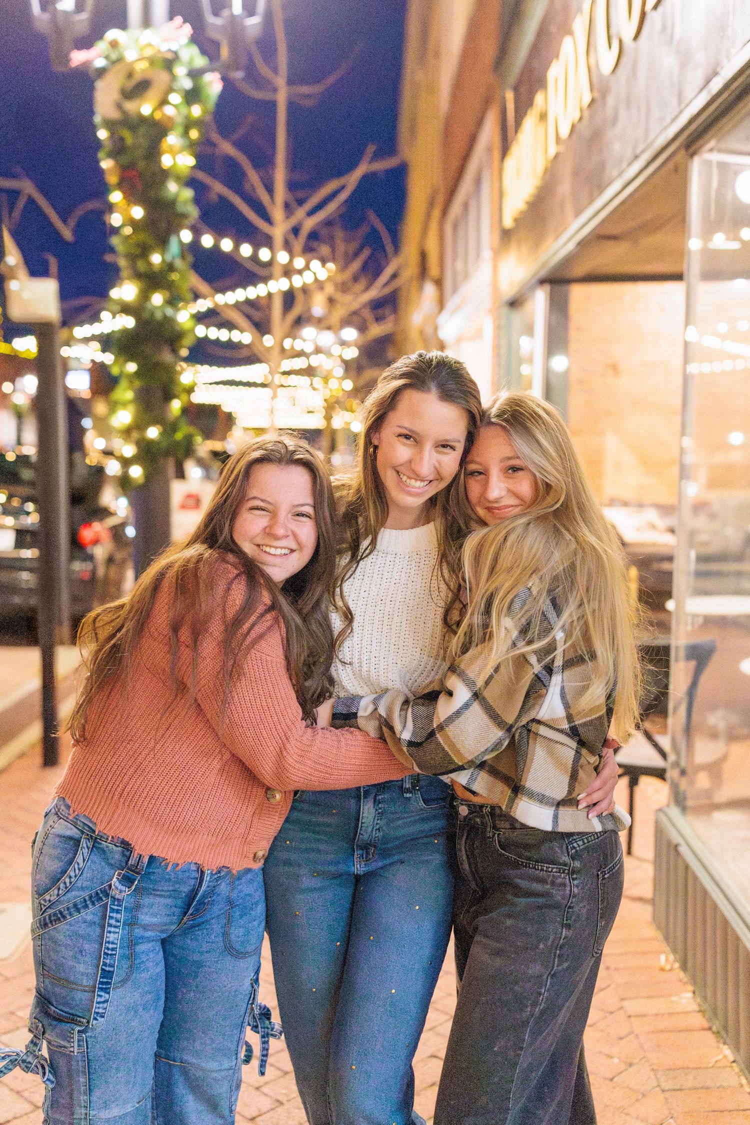 Three young women smiling and hugging on a festive lit sidewalk at night, wearing cozy sweaters and jeans.