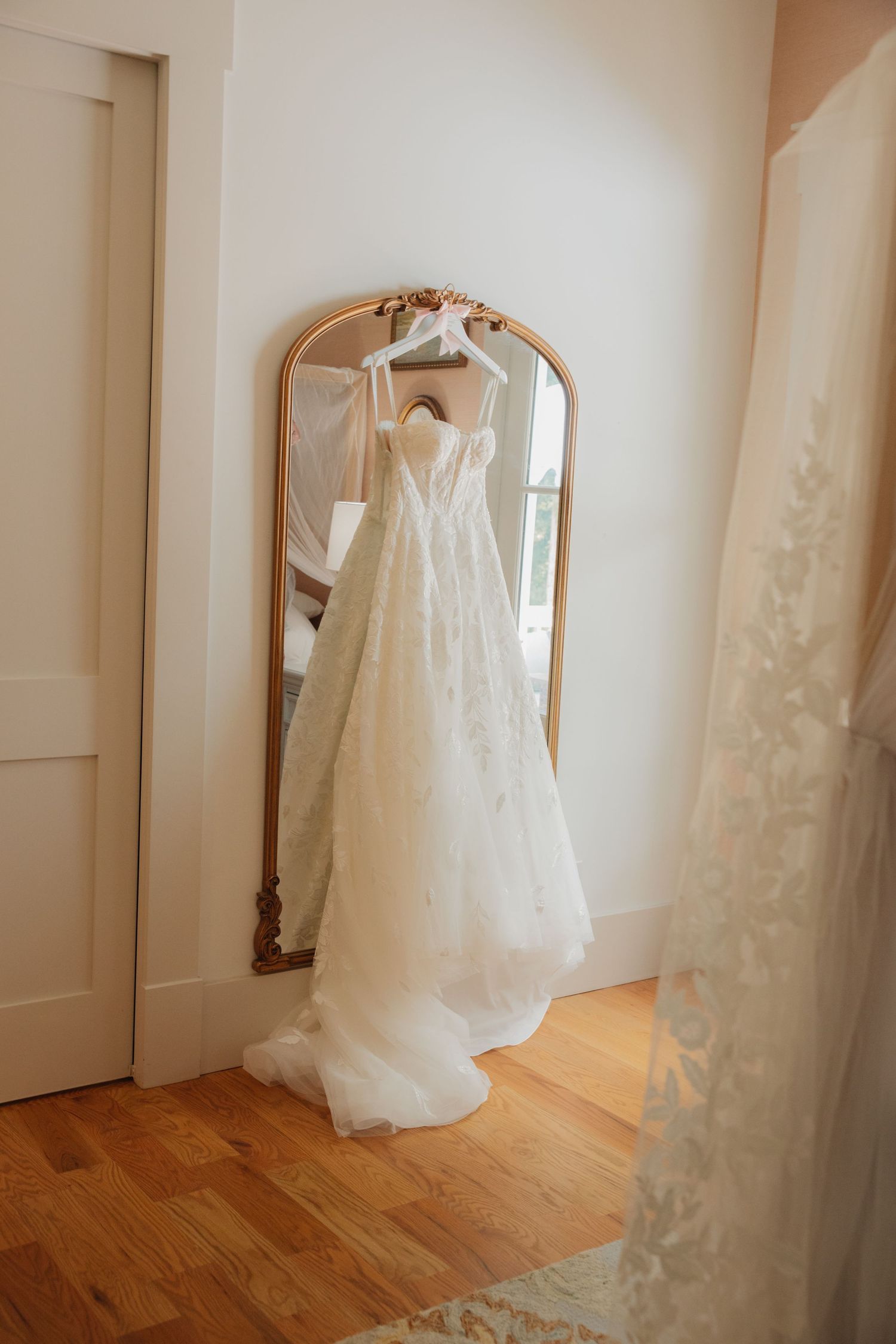 Elegant lace wedding gown hanging on a gold ornate mirror in a bright bridal suite.