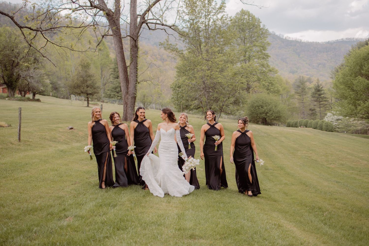 Bride in white gown poses with bridesmaids in black dresses on green lawn with mountain backdrop.