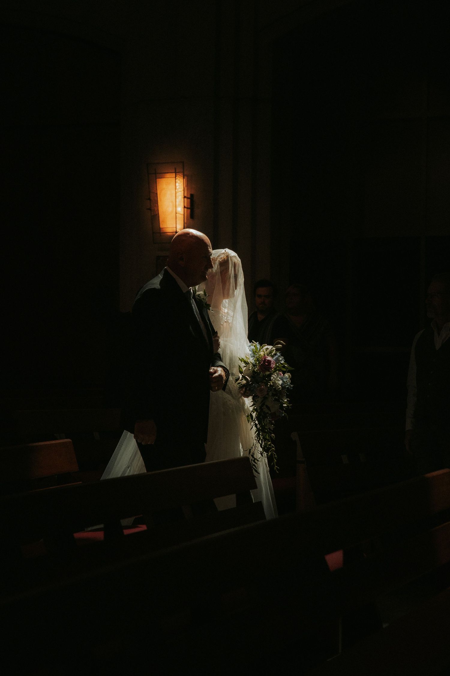 A bride and her father share a tender moment in a dimly lit church aisle, her veil and bouquet glowing softly.