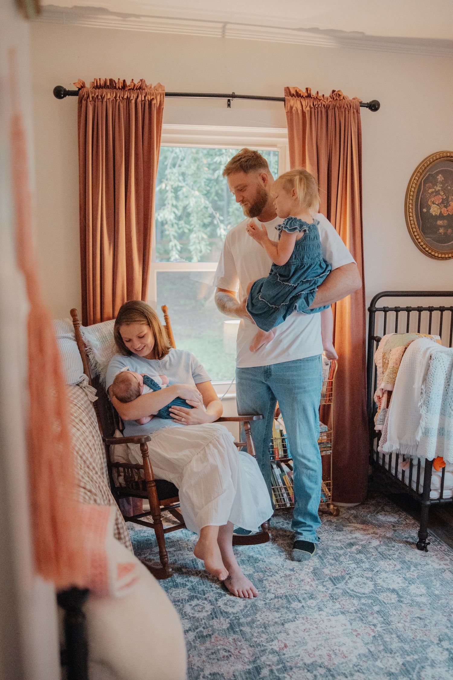 A family with a newborn baby in a cozy nursery, mother seated in rocking chair while father holds toddler by window.
