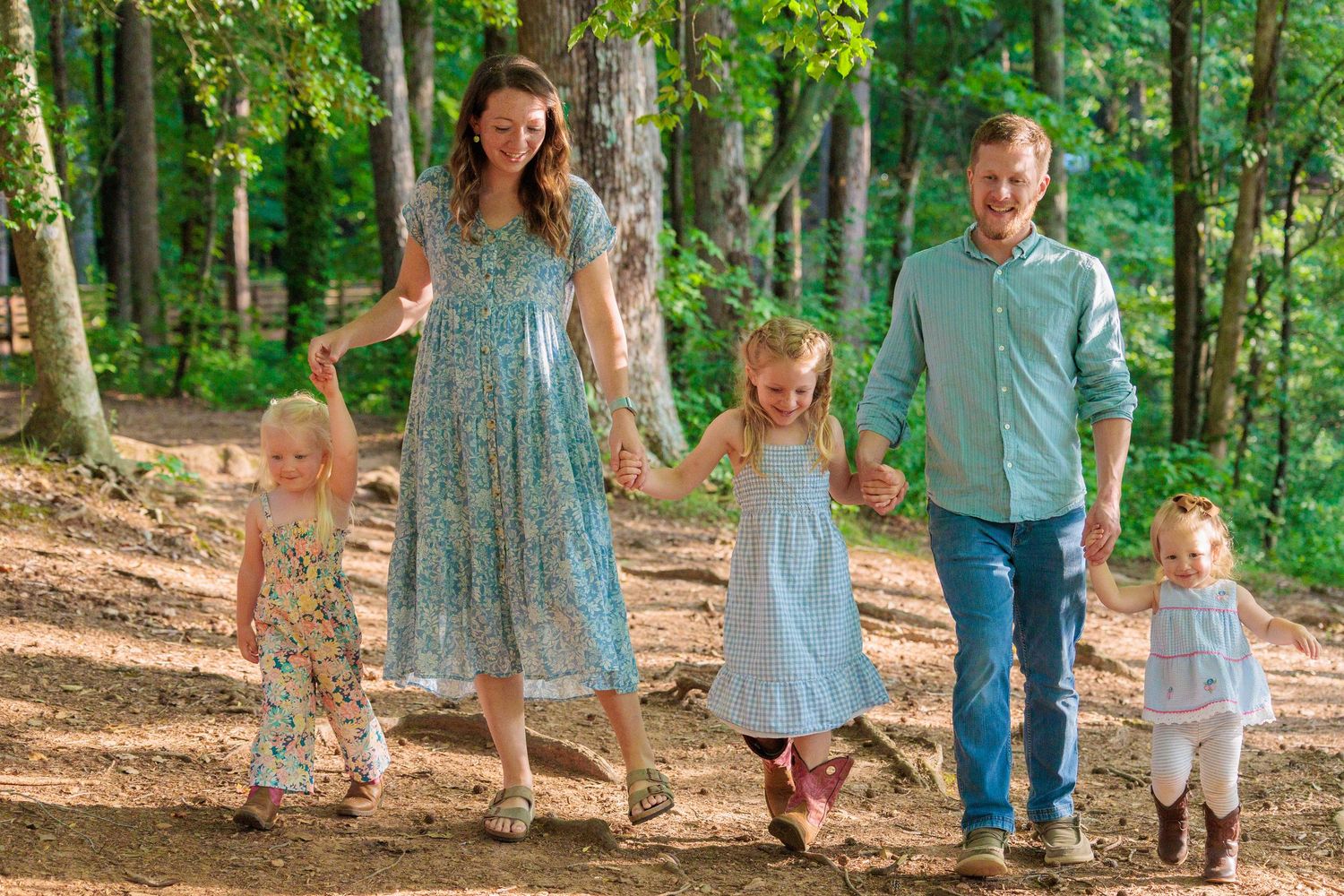 A happy family of five walks hand-in-hand through a sunlit forest path, parents and three young daughters in summer attire.