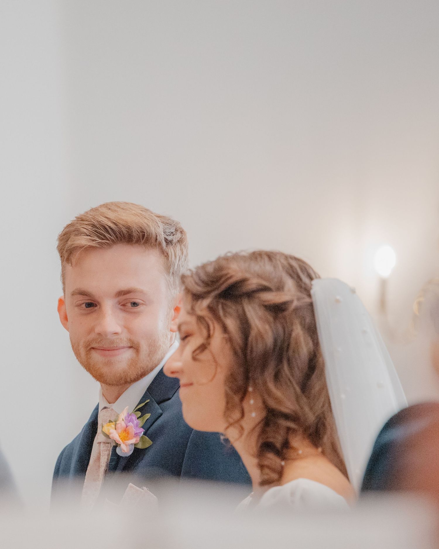 A smiling groom in a navy suit with a floral boutonniere stands beside his bride during the wedding ceremony.