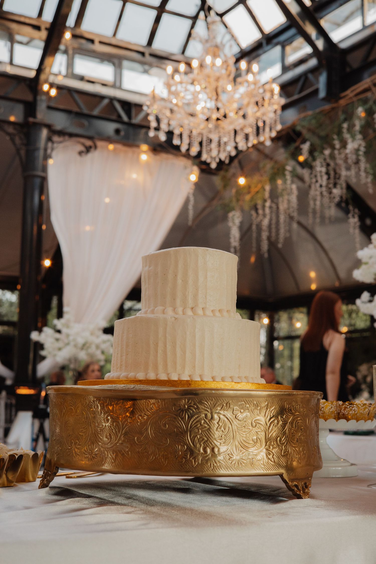 Two-tier white textured wedding cake on ornate gold stand under crystal chandelier in elegant venue.