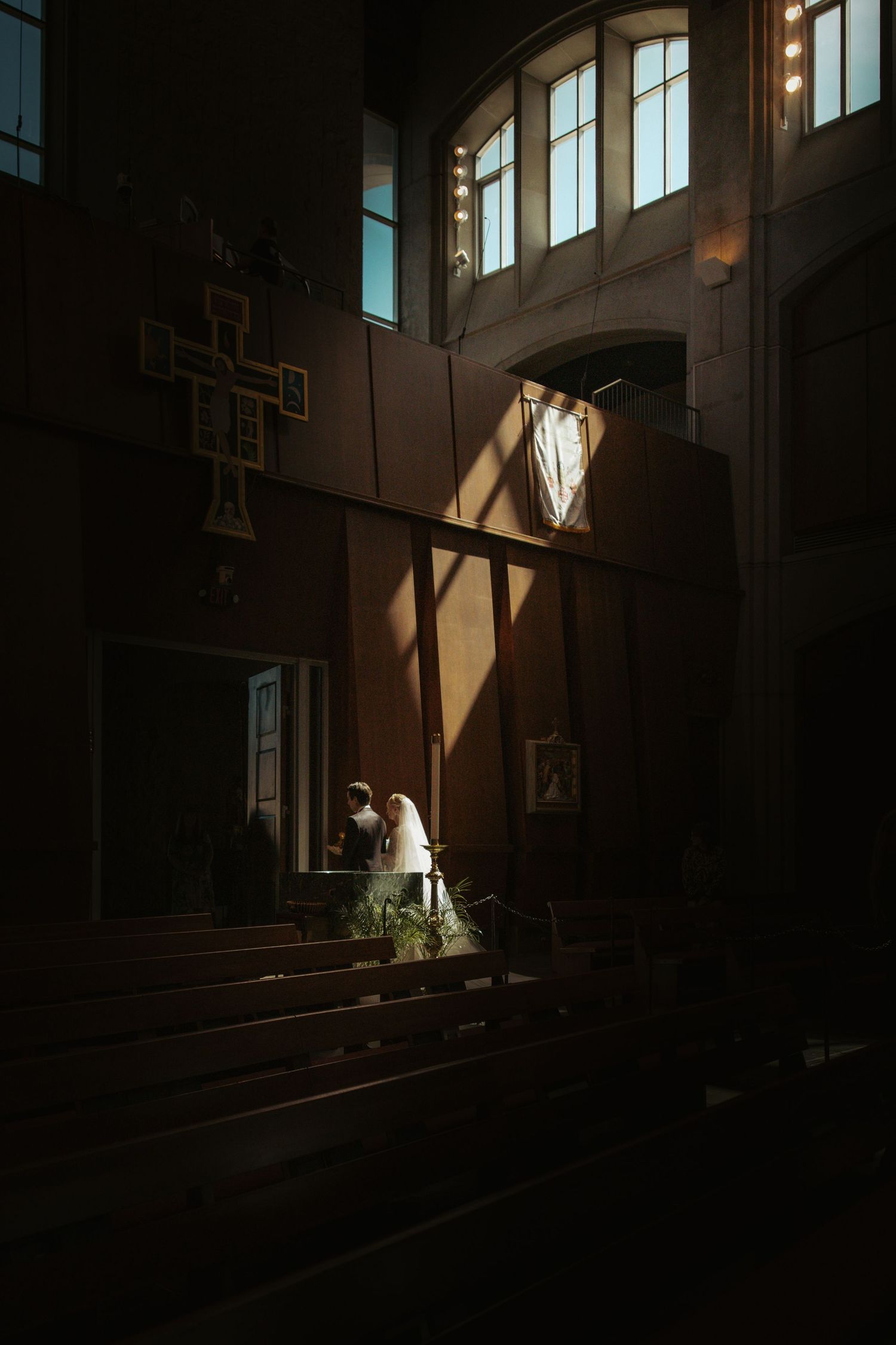 A bride and groom share a quiet moment in a dimly lit church, bathed in dramatic sunlight streaming through tall windows.