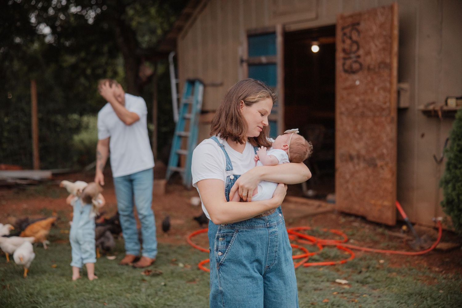 A woman in denim overalls holds a newborn baby outdoors on a farm with chickens and a rustic barn in the background.