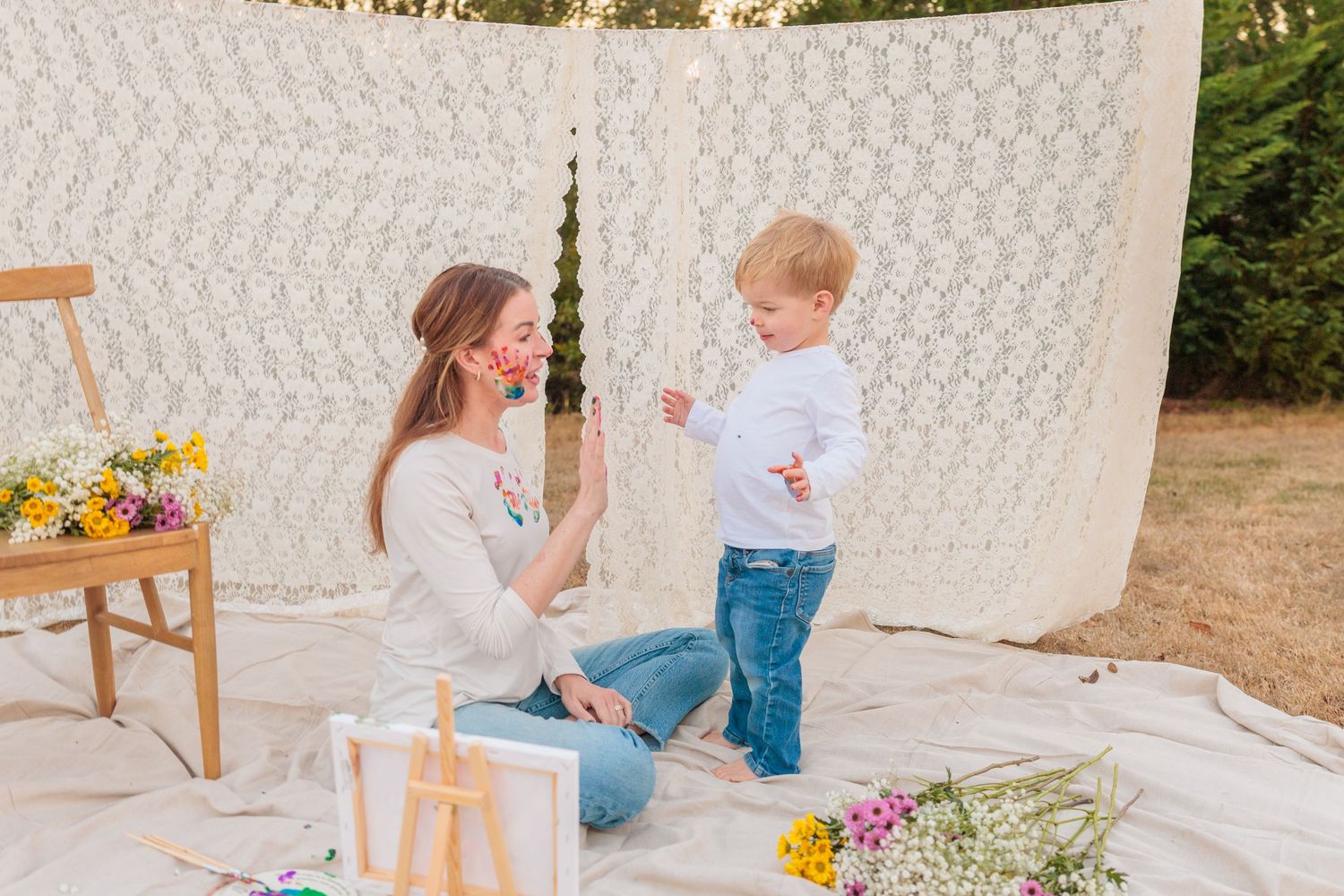 A mother and toddler play together on a blanket outdoors with a lace backdrop and colorful wildflowers nearby.