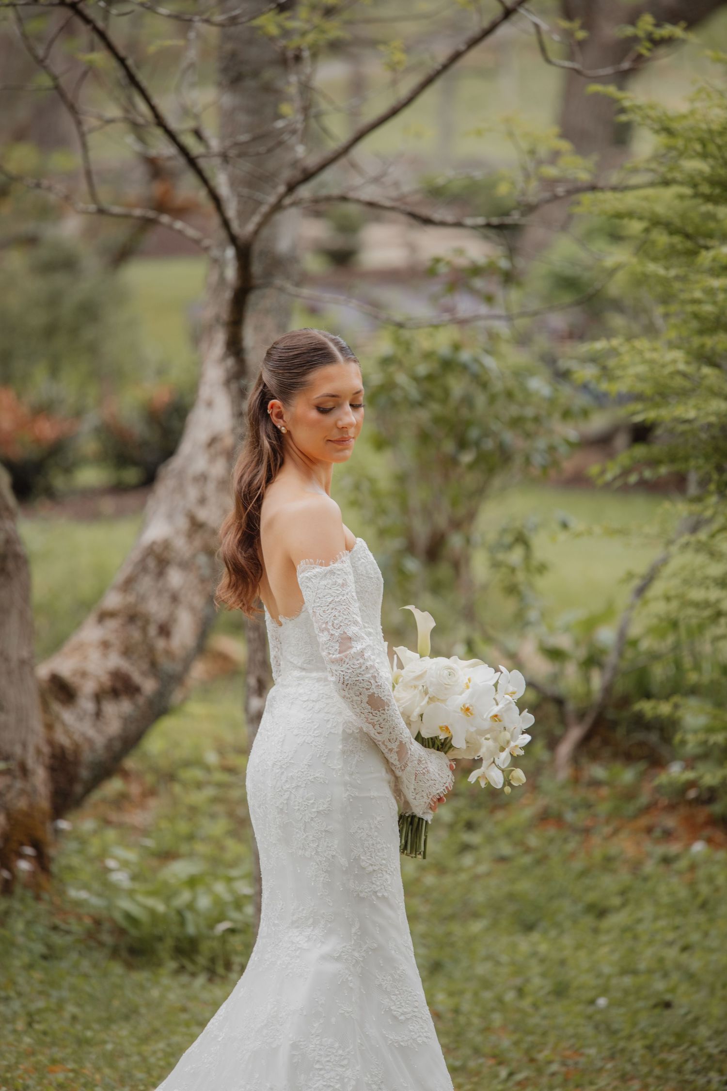 Bride in lace off-shoulder gown holding white floral bouquet in lush garden setting.