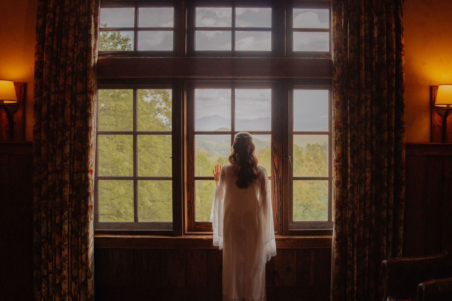 Bride in white gown stands before large wooden-framed windows, gazing at lush green trees in a warmly lit room.