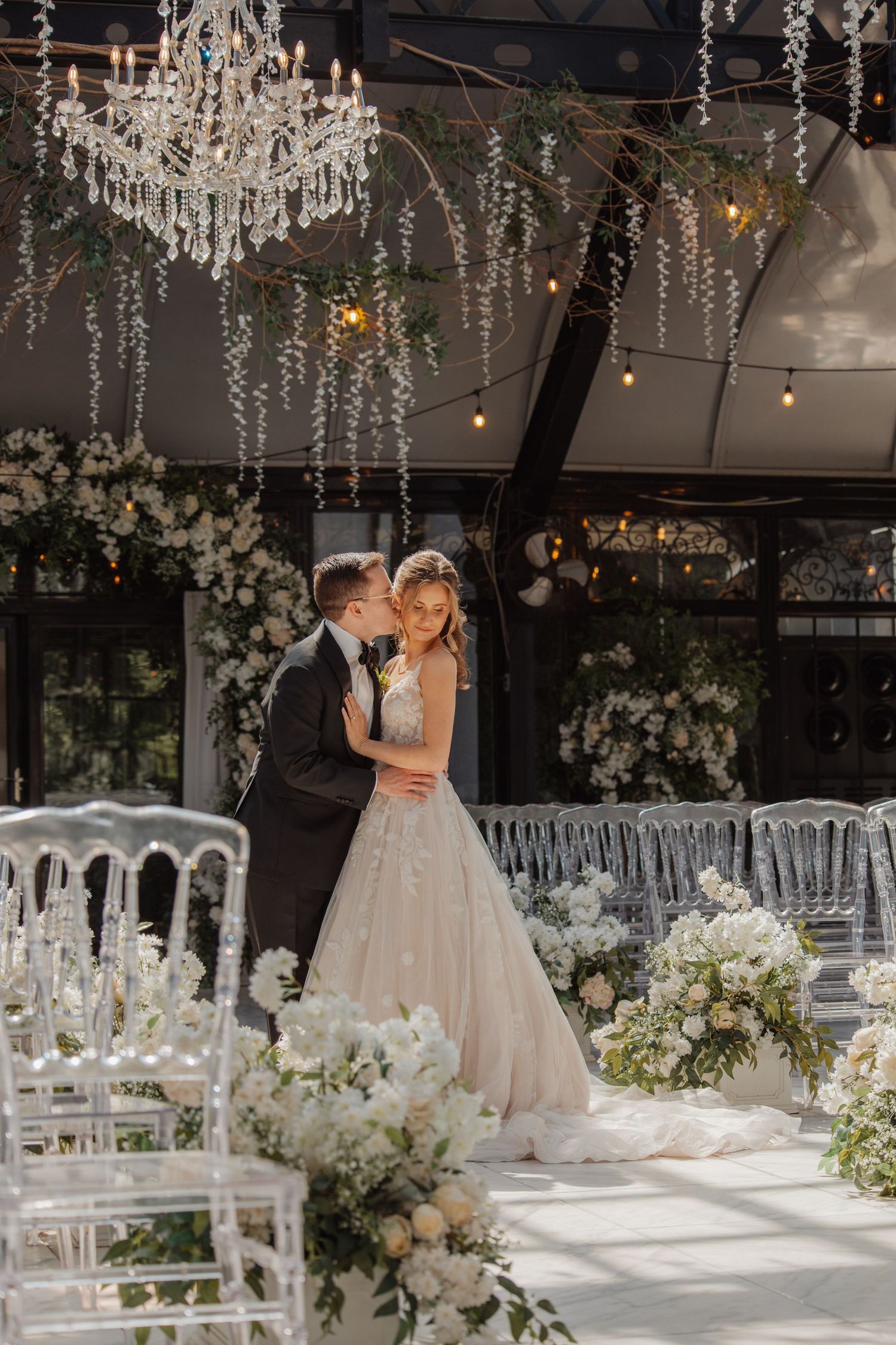 Bride and groom embracing on dance floor surrounded by white florals and crystal chandeliers.