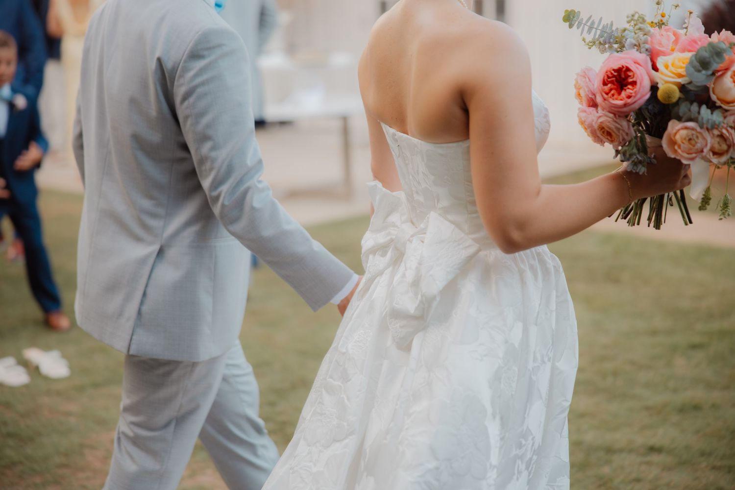Bride in strapless white gown holding pink bouquet walks hand-in-hand with groom in light blue suit.
