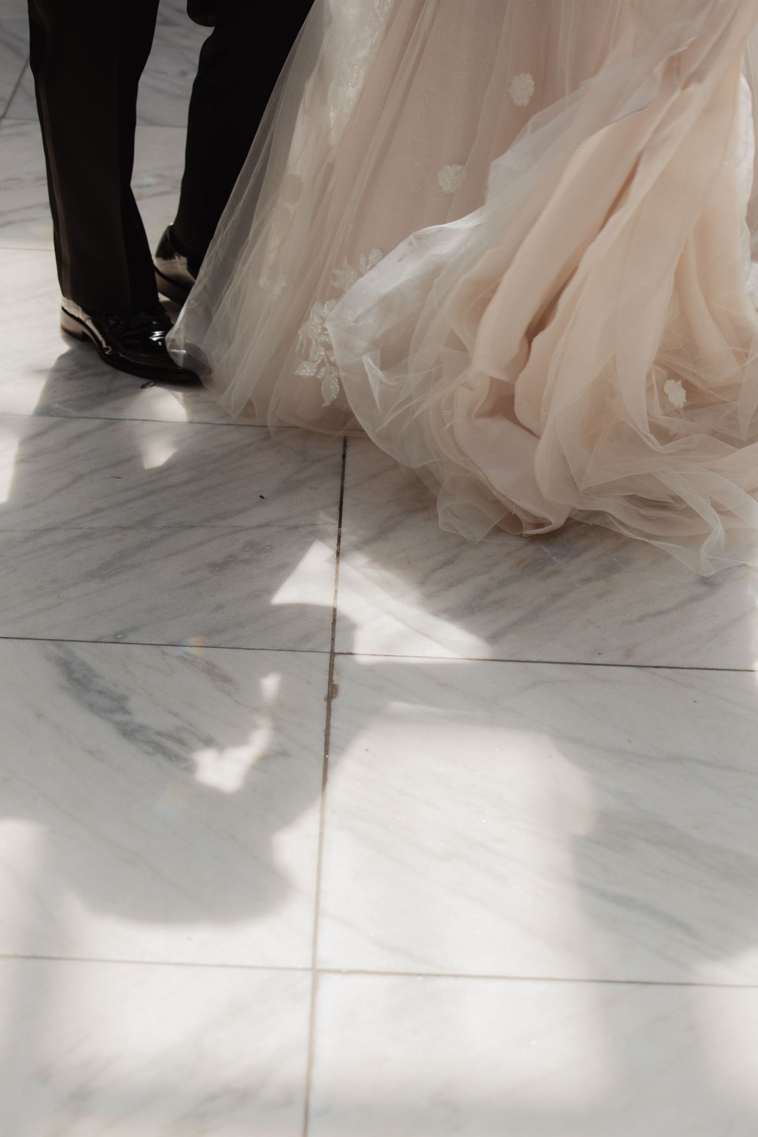 Close-up of bride's blush tulle gown and groom's dress shoes on marble floor.
