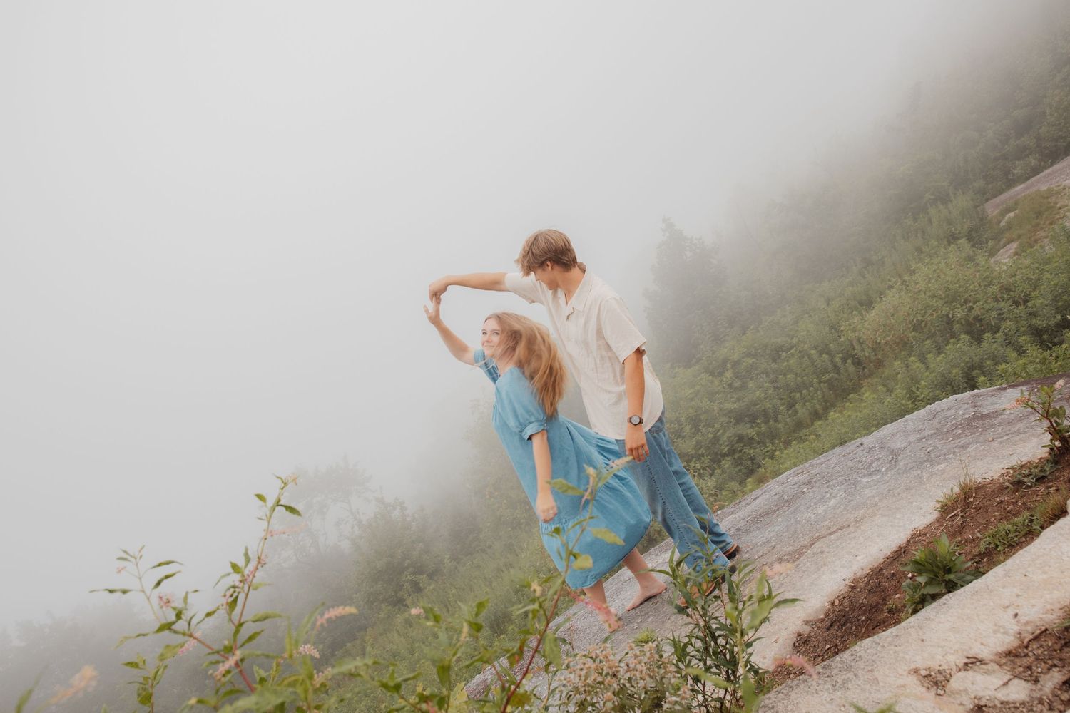 Young couple dancing playfully on a foggy rocky hillside with lush green vegetation in background.