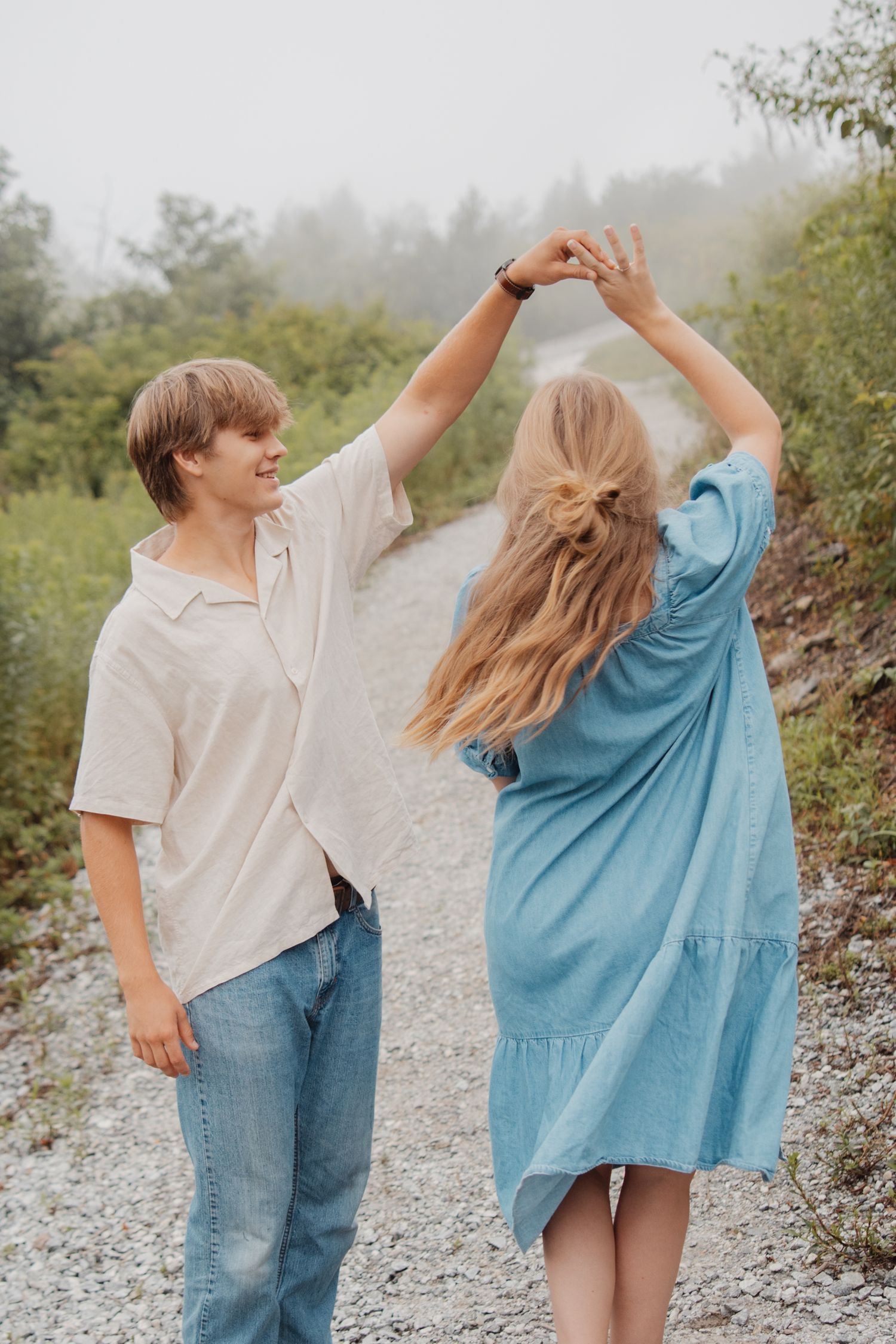 A young couple dances on a gravel path surrounded by lush greenery during an outdoor engagement session.