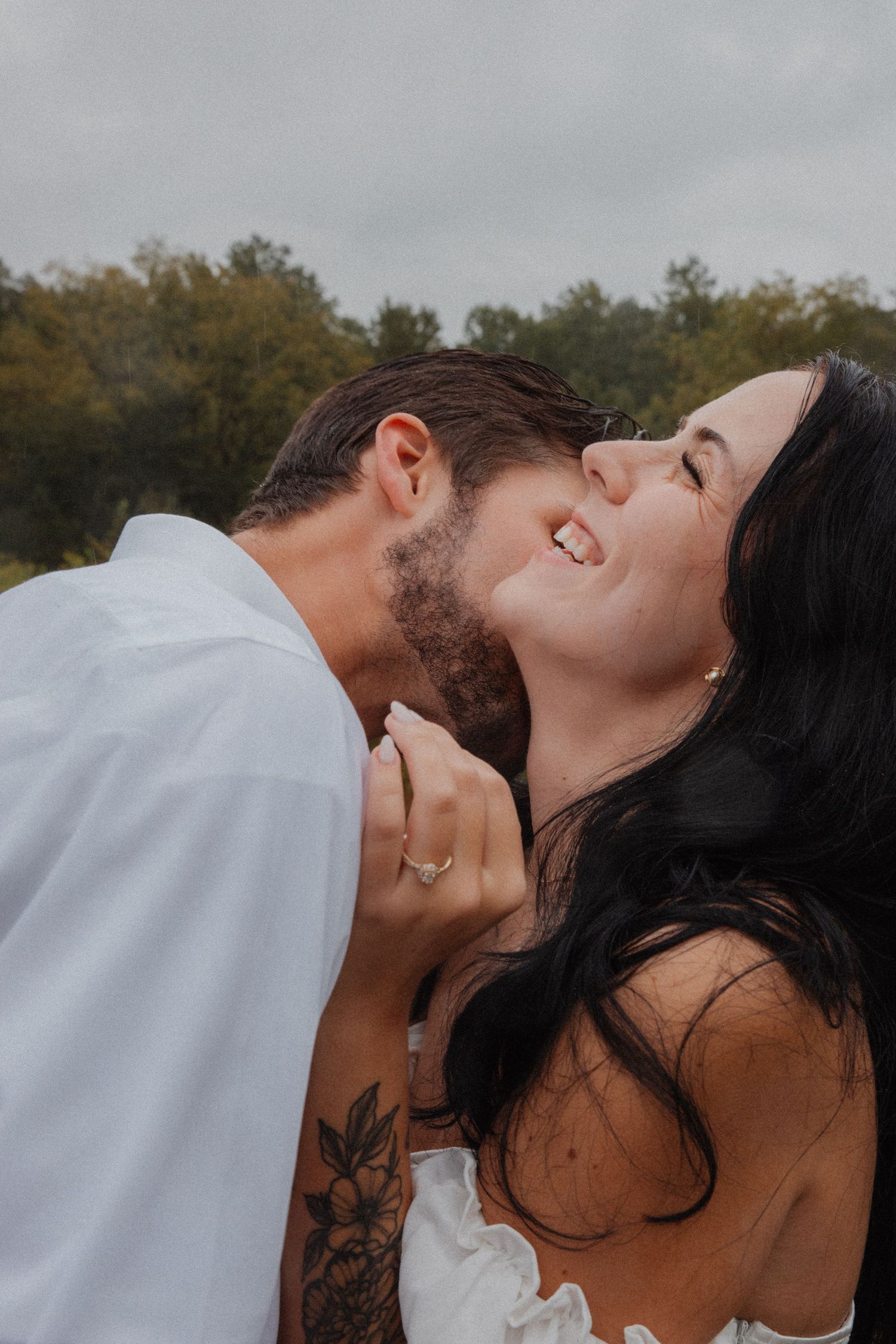 Woman laughing joyfully as man kisses her cheek outdoors under a cloudy sky.