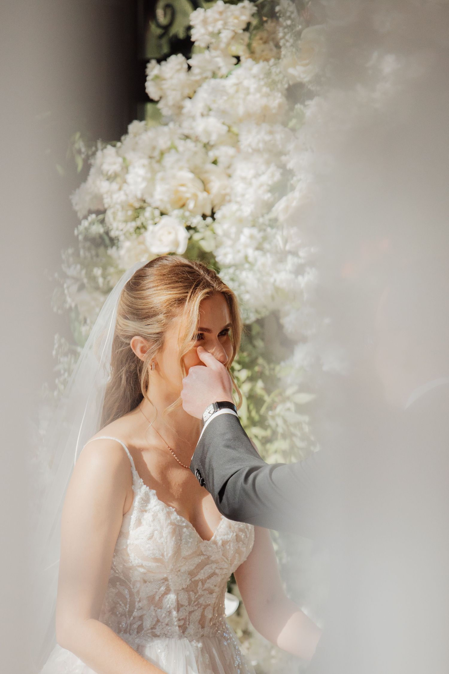 Groom gently wiping tears from bride's face during emotional wedding ceremony with white floral backdrop.
