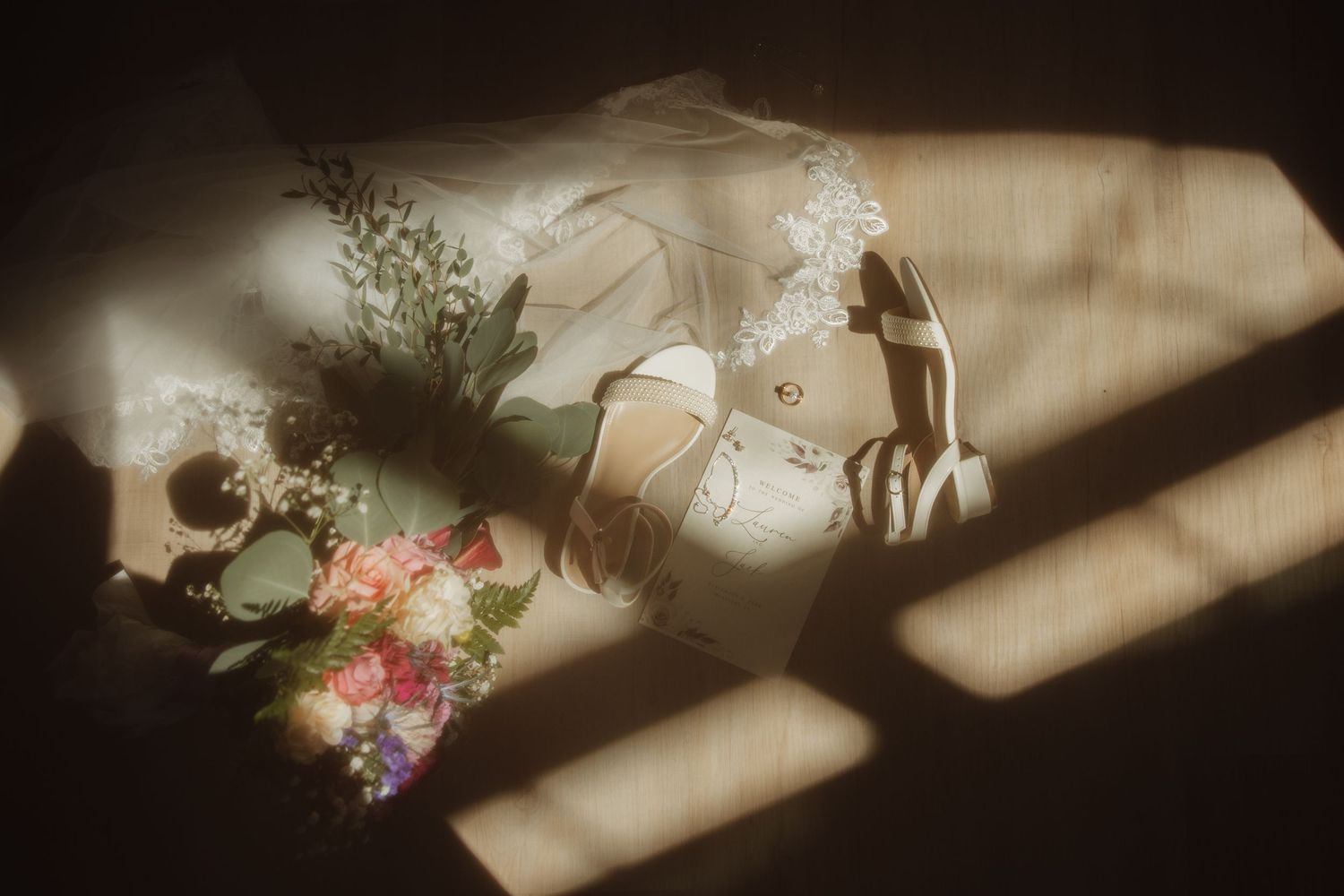 Flat lay of bridal accessories including white heels, veil, and colorful bouquet with dramatic sunlight shadows.