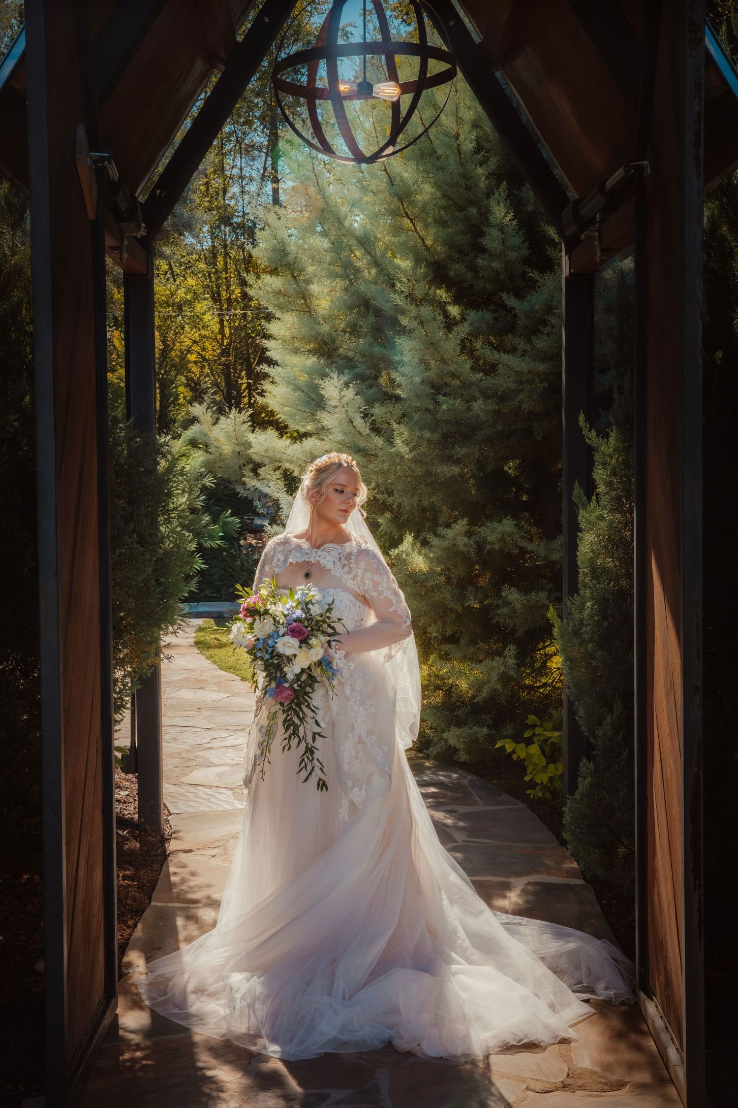 Bride in white lace gown holding floral bouquet stands in elegant wooden archway surrounded by lush greenery.