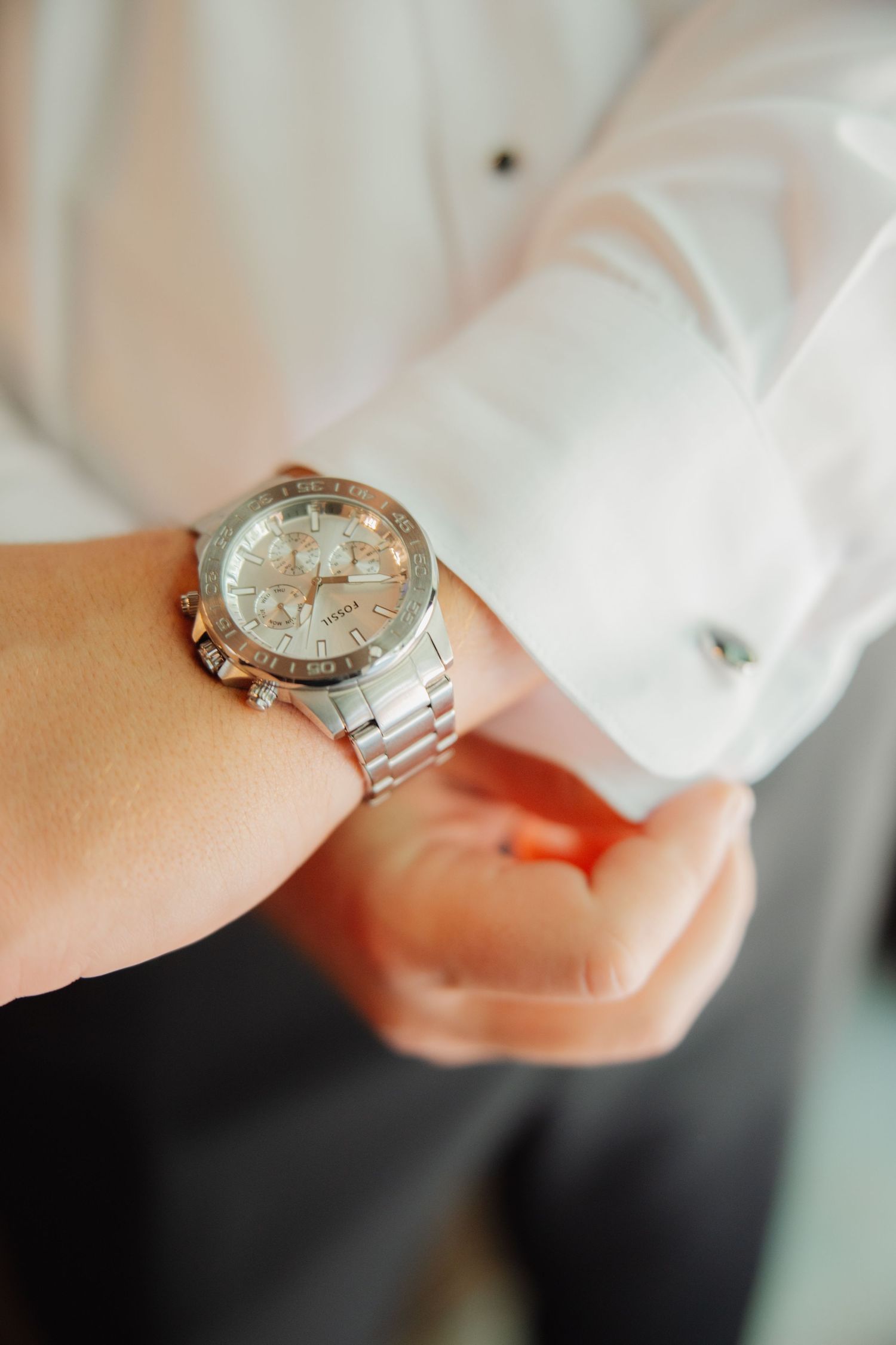 Groom in white dress shirt adjusting silver chronograph watch with cufflinks on wedding day.