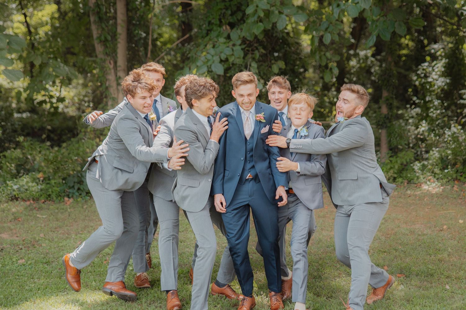 Groom in navy suit playfully surrounded by groomsmen in gray suits laughing outdoors at wedding celebration.