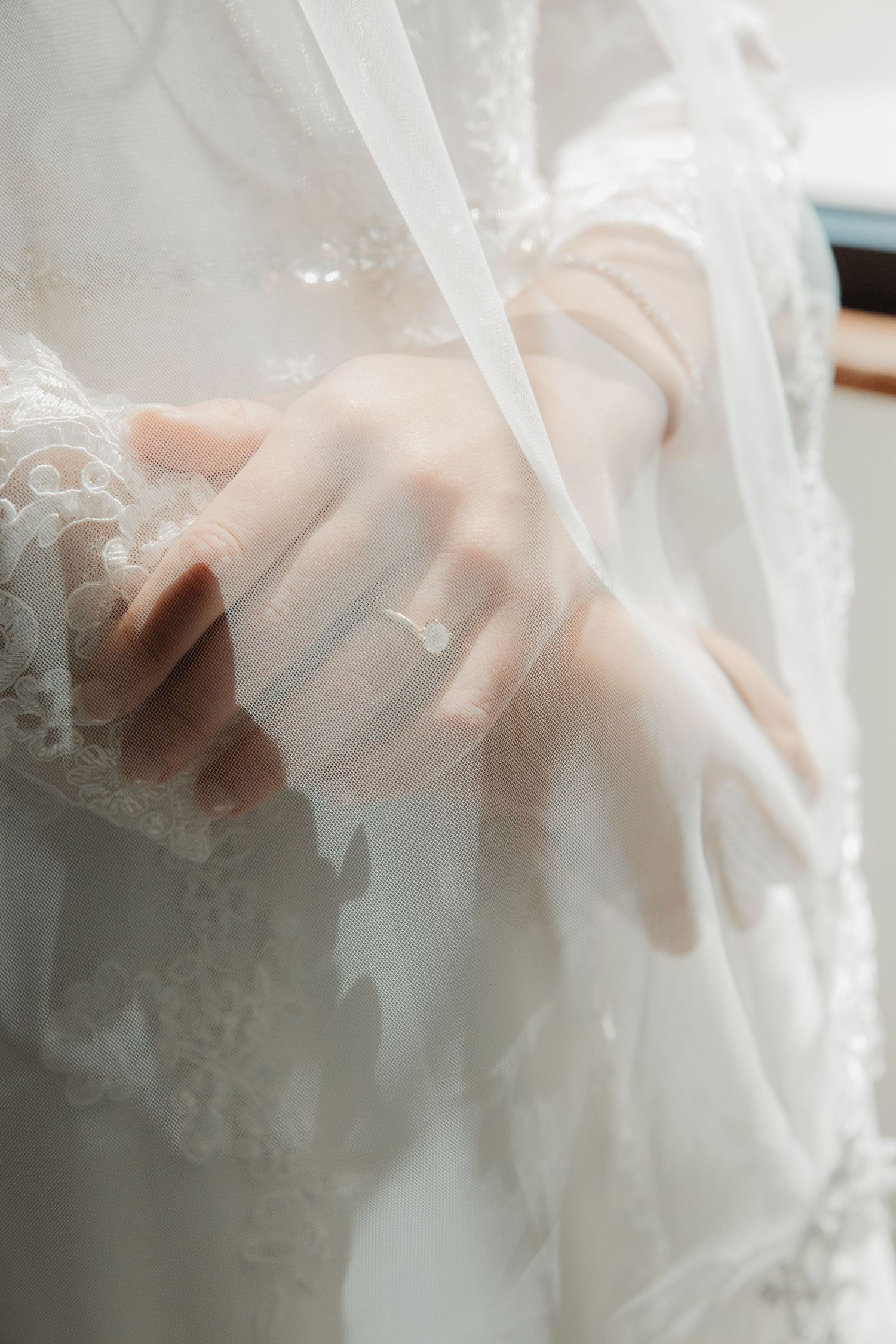 Close-up of bride's hands folded over white lace wedding dress with delicate diamond engagement ring.
