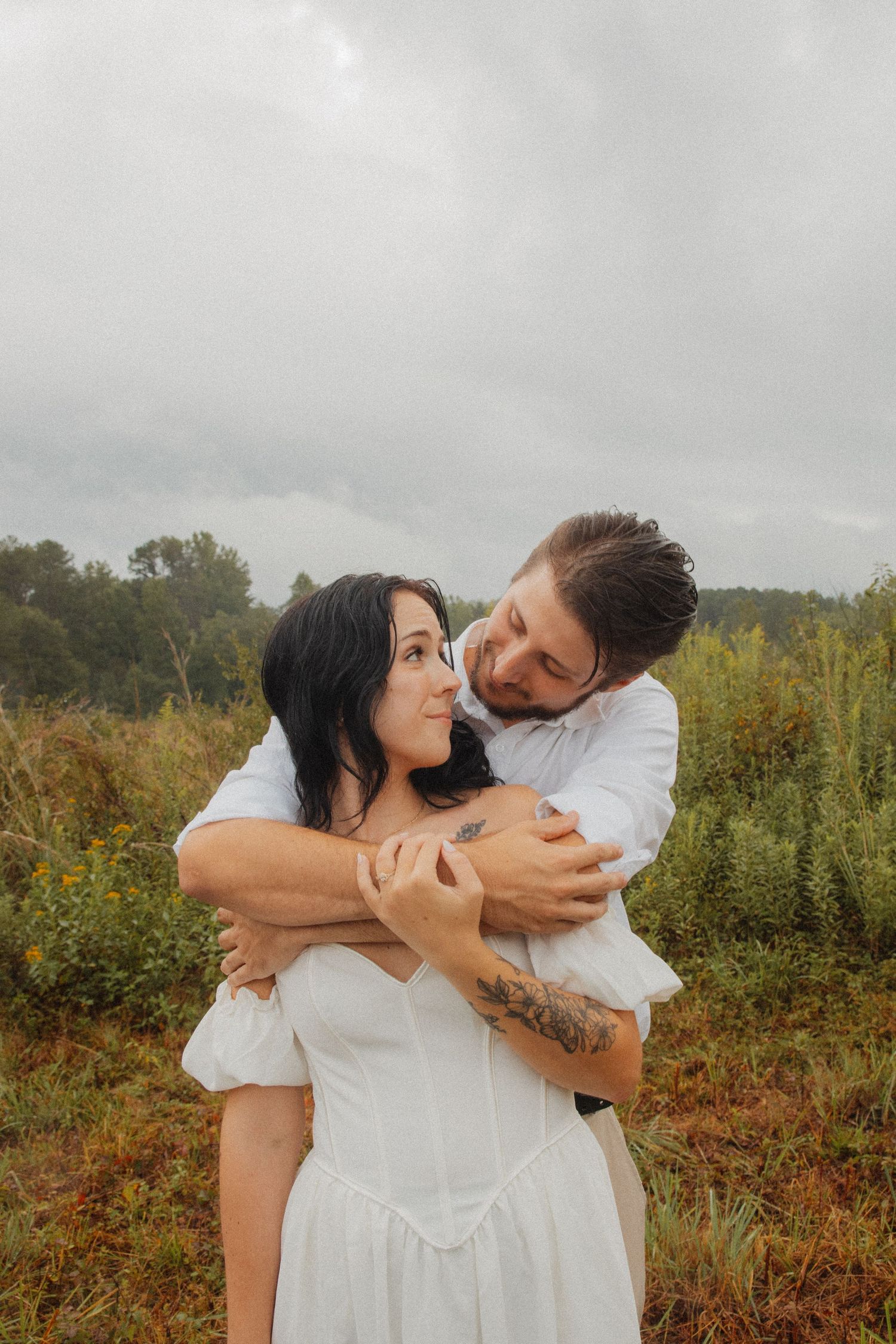 Man hugging woman from behind in a green field under an overcast sky.