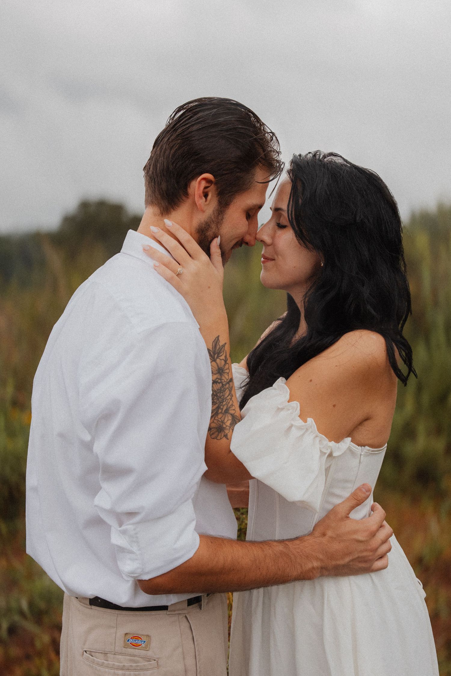 Romantic couple in white outfits embracing nose-to-nose in a lush green field on cloudy day.