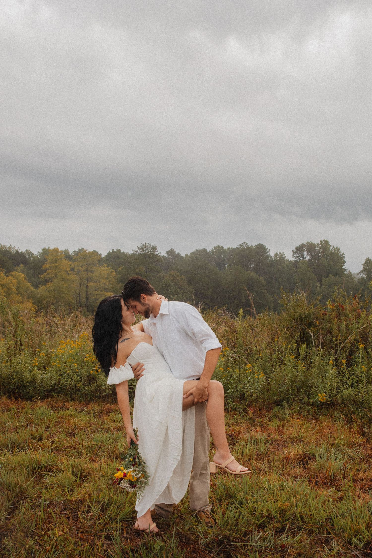 Couple sharing a romantic kiss in a wildflower meadow under a cloudy sky.